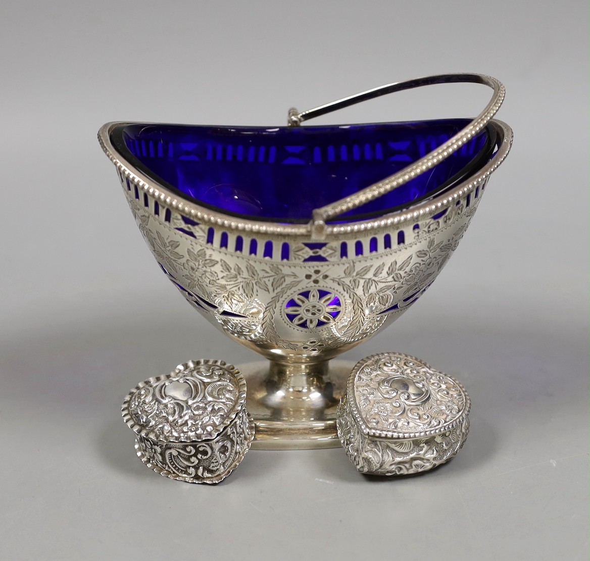 A late Victorian pierced silver sugar basket, by Nathan & Hayes, Chester, 1896, with blue glass liner, length 13cm, together with two repousse silver heart shaped pill boxes.